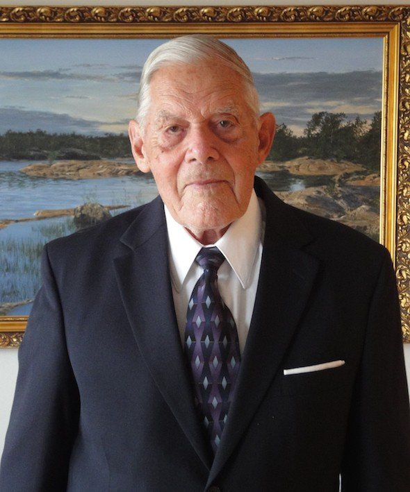 Photograph of an older man in a dark suit standing before a framed painting
