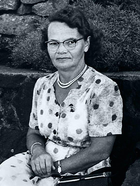 Black-and-white photograph of a woman in a polka-dot dress seated by a stone wall